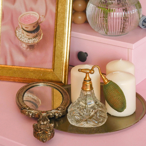 Coquette bridal shower theme inspiration image of vintage mirror, perfume bottle, candles, teacups and glass vase on a pink dresser.