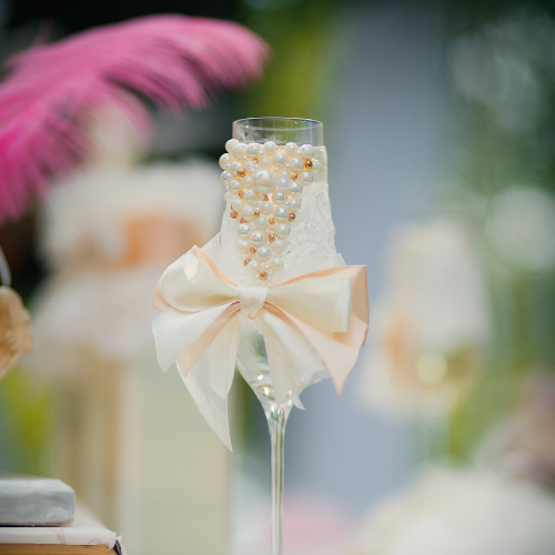 Coquette bridal shower theme inspiration image of a wine glass decorate with pearls a silky cream blush bow and lace and a pink feather in the background.