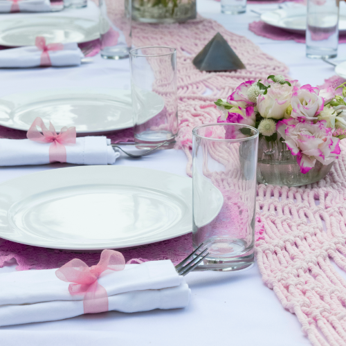 Coquette bridal shower theme inspiration image of an event table setting with boho macrame table runner, white plates and napkins with pink organza bows.