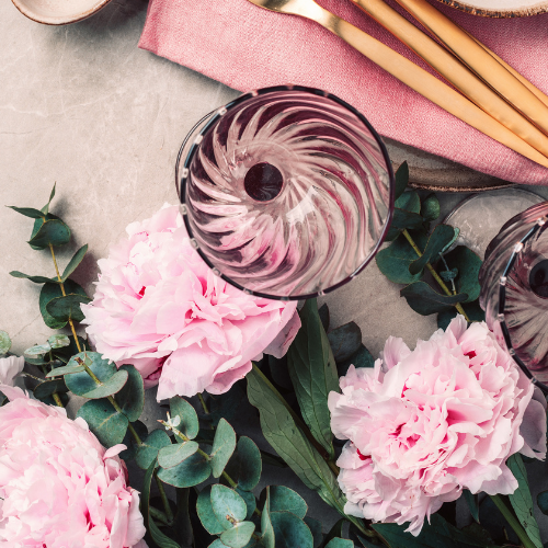 Coquette bridal shower theme inspiration image of tinted glass swirly wine glasses, gold cutlery and pink peony flowers with eucalyptus foliage.